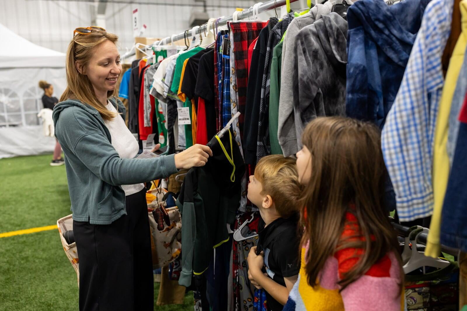 Woman shopping secondhand clothing with her young children looking on.